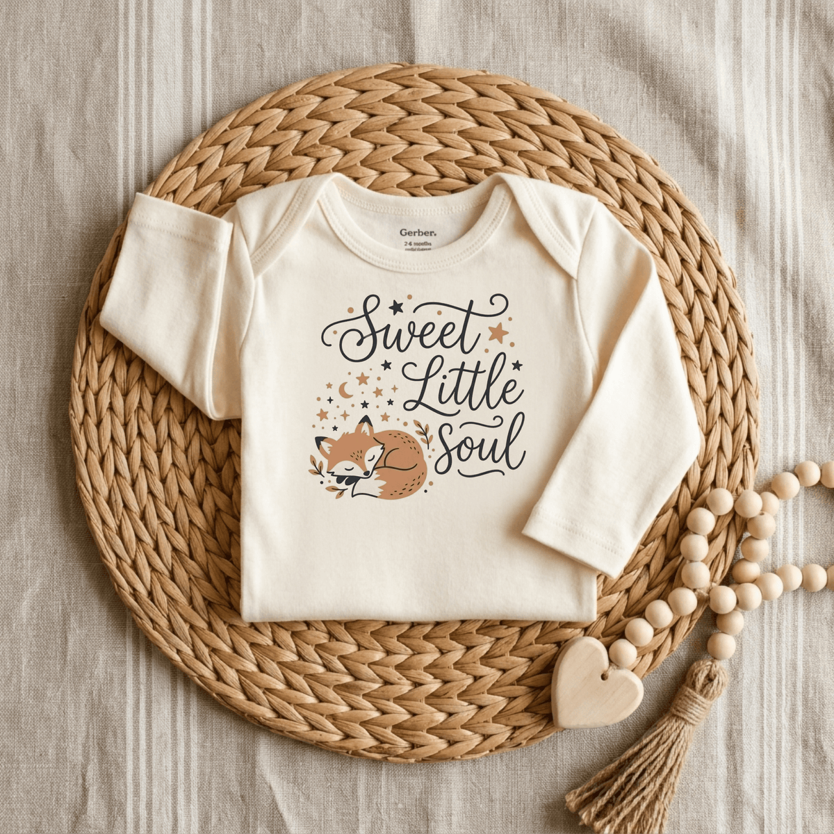 A Toodleroos Sweet Little Soul Onesie – Cute Sleeping Fox Bodysuit in a creamy shade rests on a woven mat beside a wooden bead and heart garland, all arranged over a striped fabric background.