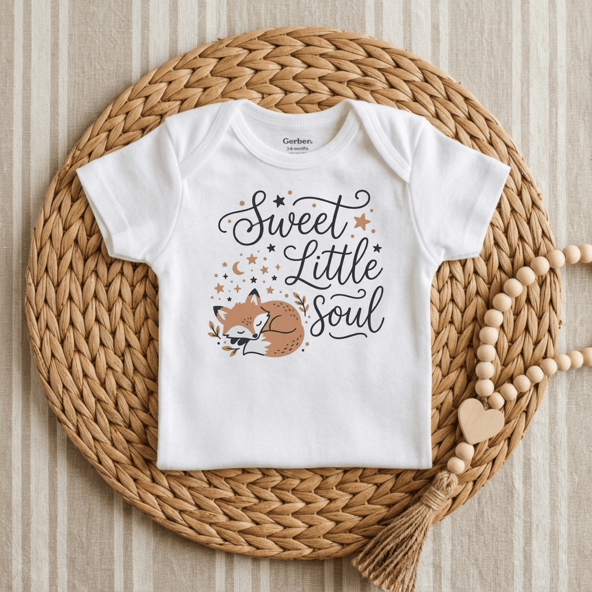 A Toodleroos Sweet Little Soul Onesie – Cute Sleeping Fox Bodysuit is displayed on a woven mat, featuring a gender-neutral white design with a sleeping fox, stars, and the phrase “Sweet Little Soul.” A beaded wooden garland with a heart and tassel lies beside this charming woodland baby bodysuit.