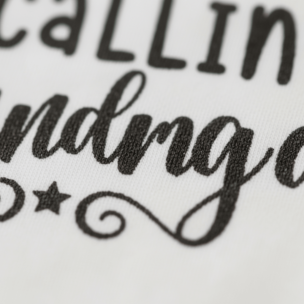 Close-up of black, decorative text and a star with a swirl on white fabric from the That’s It I’m Calling Grandma Baby Onesie by Toodleroos. The bold and elegant partially visible text makes this funny bodysuit a perfect coming-home outfit or thoughtful baby shower gift.