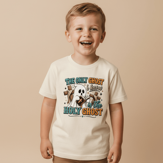 A smiling young boy wears the Toodleroos “The Only Ghost I Know Kids Shirt – Holy Ghost Halloween Tee,” featuring a cartoon ghost and the phrase, “The only ghost I know is the Holy Ghost,” set against a plain beige background—a faith-inspired Halloween shirt perfect for little believers.