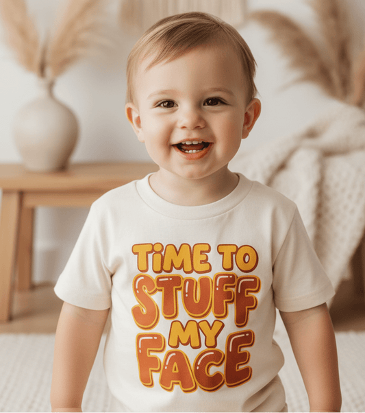 A smiling toddler wears the Toodleroos “Time to Stuff My Face Thanksgiving Toddler Shirt”—a funny Turkey Day tee in a cream color, featuring bold, playful lettering—standing indoors surrounded by neutral decor and dried plants, perfect for holiday kids clothing.