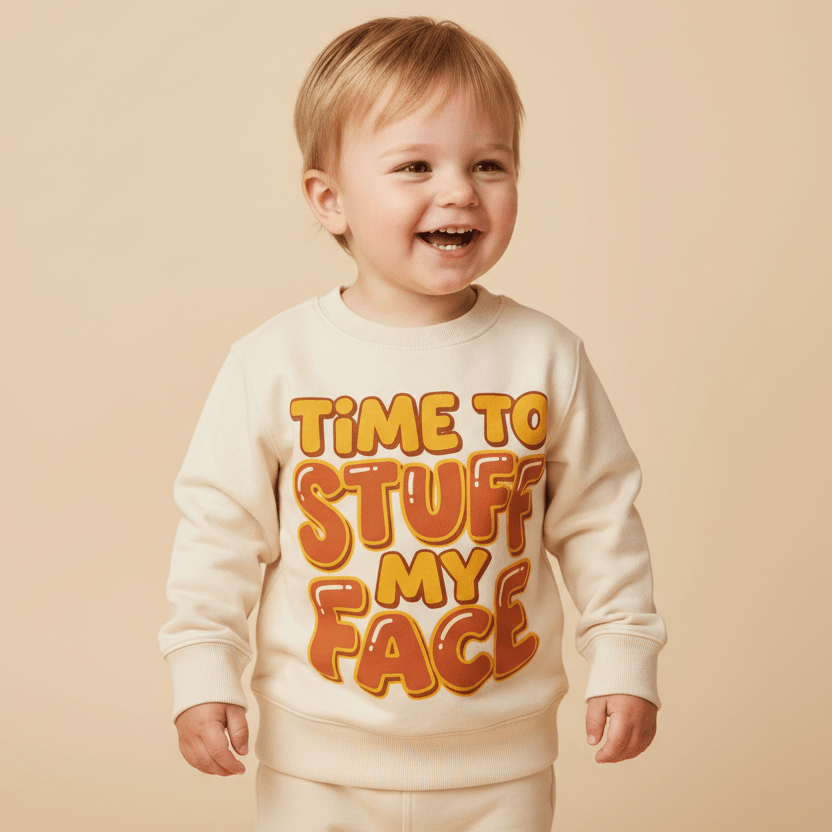 A smiling toddler wears the Toodleroos “Time to Stuff My Face” Thanksgiving Toddler Sweatshirt—a funny Turkey Day pullover featuring bold orange and yellow lettering—while standing against a beige background.