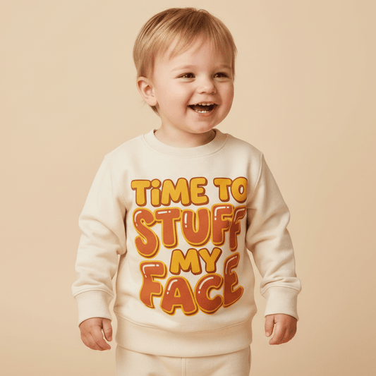 A smiling toddler wears the Toodleroos “Time to Stuff My Face” Thanksgiving Toddler Sweatshirt—a funny Turkey Day pullover featuring bold orange and yellow lettering—while standing against a beige background.