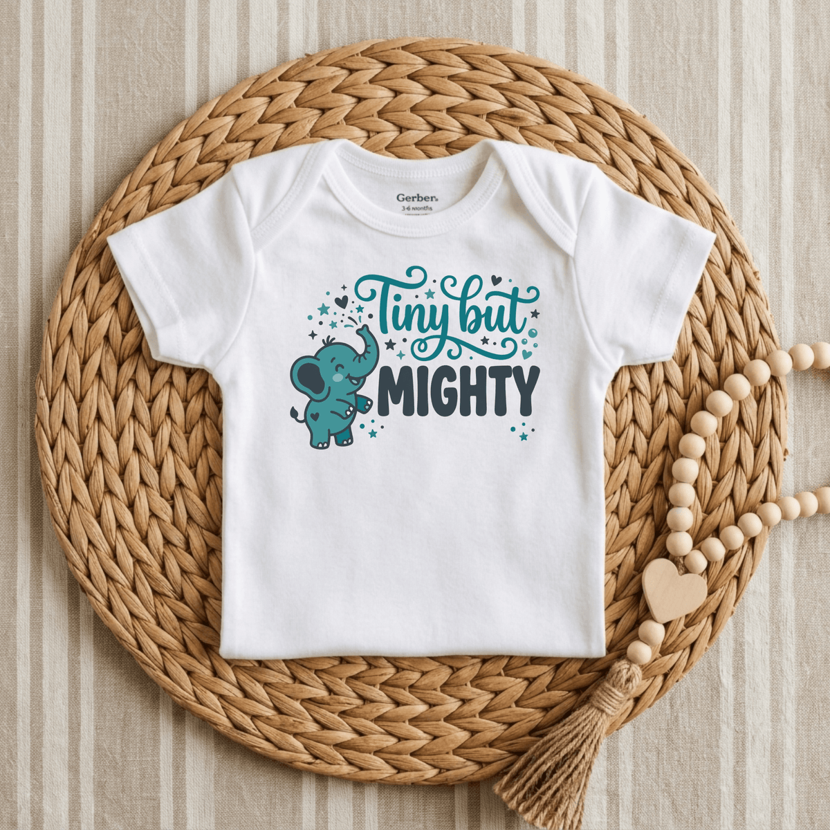 The Toodleroos Tiny But Mighty Onesie – Cute Baby Elephant Bodysuit features an adorable blue elephant and teal stars graphic with the phrase “Tiny but Mighty.” This gender-neutral onesie is a thoughtful preemie baby gift or NICU graduate outfit. Displayed on a woven mat beside a wooden bead garland with a heart, it makes for a charming presentation.
