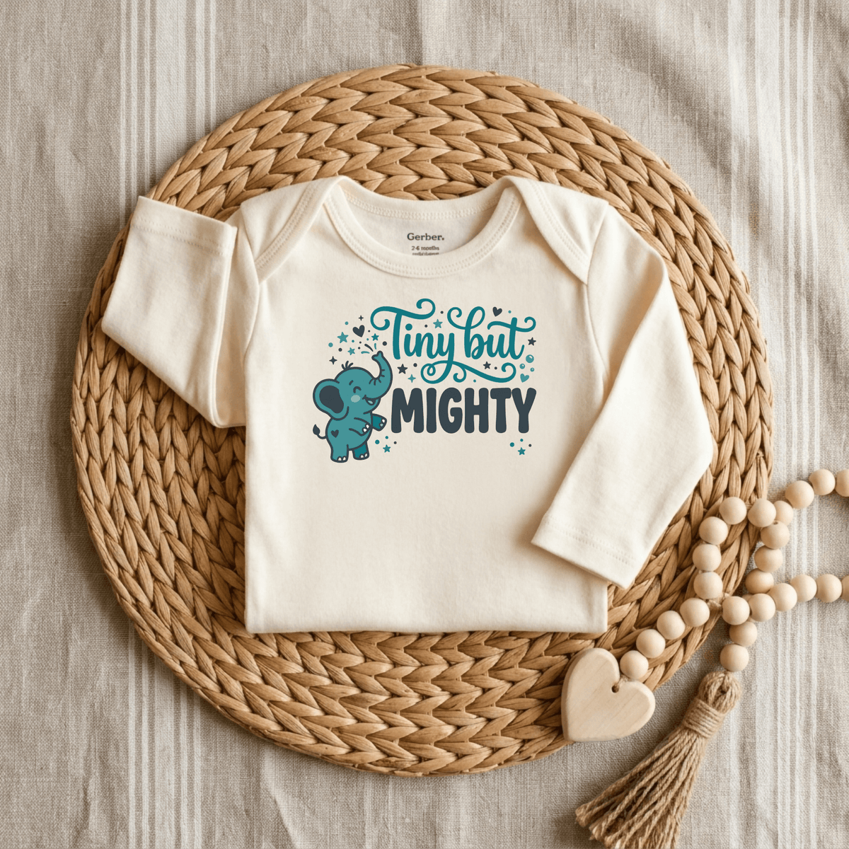 Showcase your little one in the Toodleroos Tiny But Mighty Onesie – a cute, cream-colored, long-sleeve baby bodysuit featuring a blue cartoon elephant and bold “Tiny but MIGHTY” text. Styled on a round wicker placemat with decorative wooden beads against a textured linen backdrop, this gender-neutral outfit makes an ideal preemie baby gift or a special way to celebrate your NICU graduate.