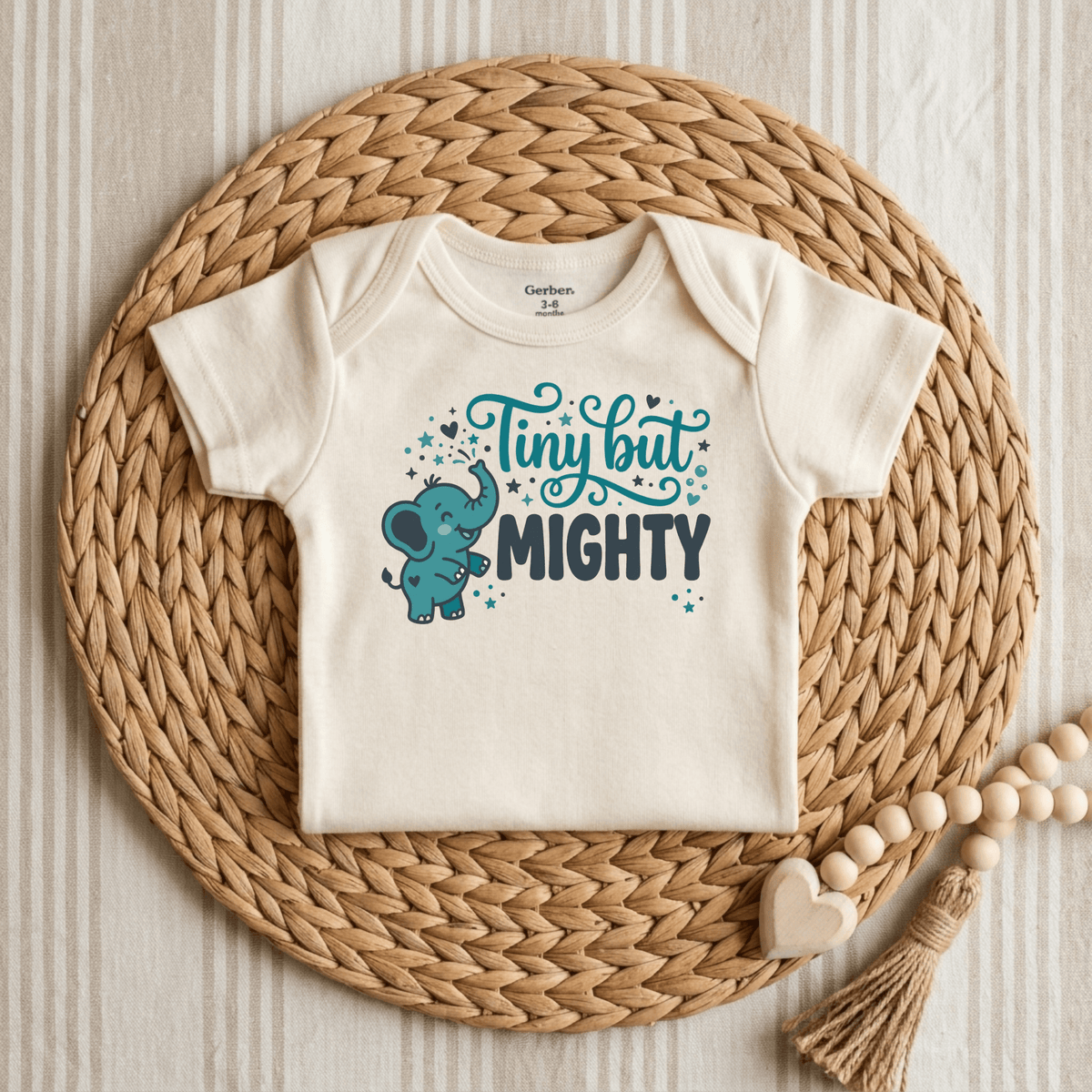 Celebrate your little NICU graduate with the Toodleroos Tiny But Mighty Onesie – a gender-neutral bodysuit featuring a charming blue elephant and the phrase “Tiny but Mighty.” Presented on a woven placemat next to a wooden rattle and set against a striped cloth background, it’s the perfect preemie baby gift.