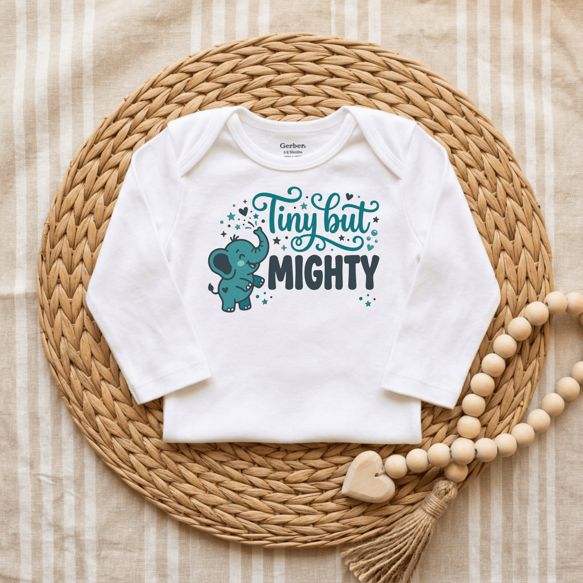 The Toodleroos Tiny But Mighty Onesie – Cute Baby Elephant Bodysuit is a white, gender-neutral baby onesie featuring an adorable blue elephant illustration and the words “Tiny but MIGHTY.” Displayed on a woven mat beside a wooden beaded garland with a heart charm, it’s an ideal preemie baby gift or NICU graduate onesie.