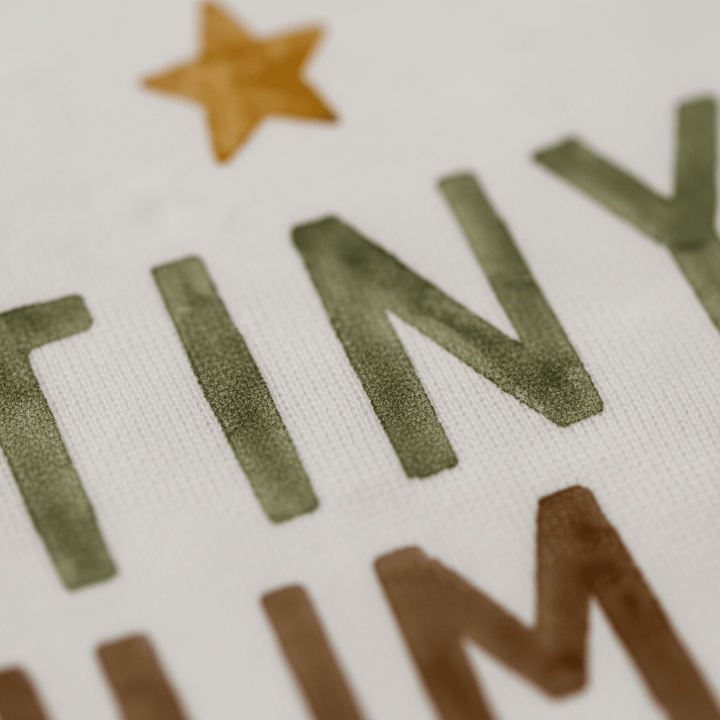 Close-up of fabric featuring the word “TINY” in green letters, part of a gold star, and brown text above and below, showcasing the textured minimalist design of the Toodleroos Gender-Neutral Baby Onesie – Tiny Human Short-Sleeve Graphic Bodysuit.