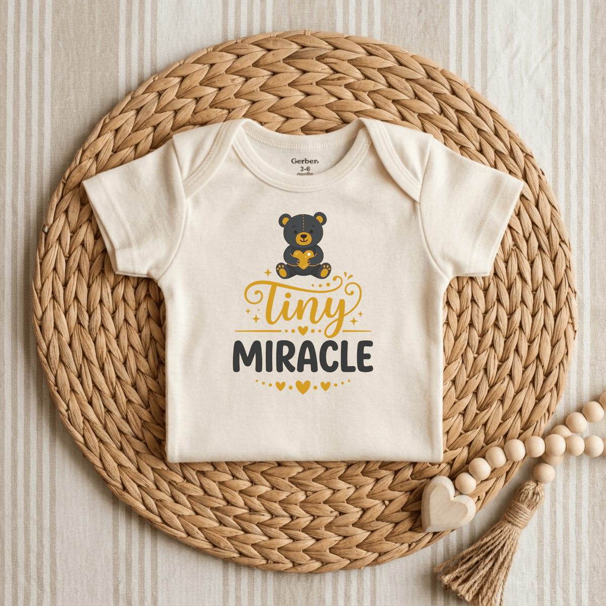 The Tiny Miracle Onesie by Toodleroos features a charming teddy bear graphic and the phrase “tiny MIRACLE” in yellow and black. Placed on a woven mat next to a sweet wooden beaded heart toy, this adorable newborn bodysuit makes a thoughtful NICU graduate gift.