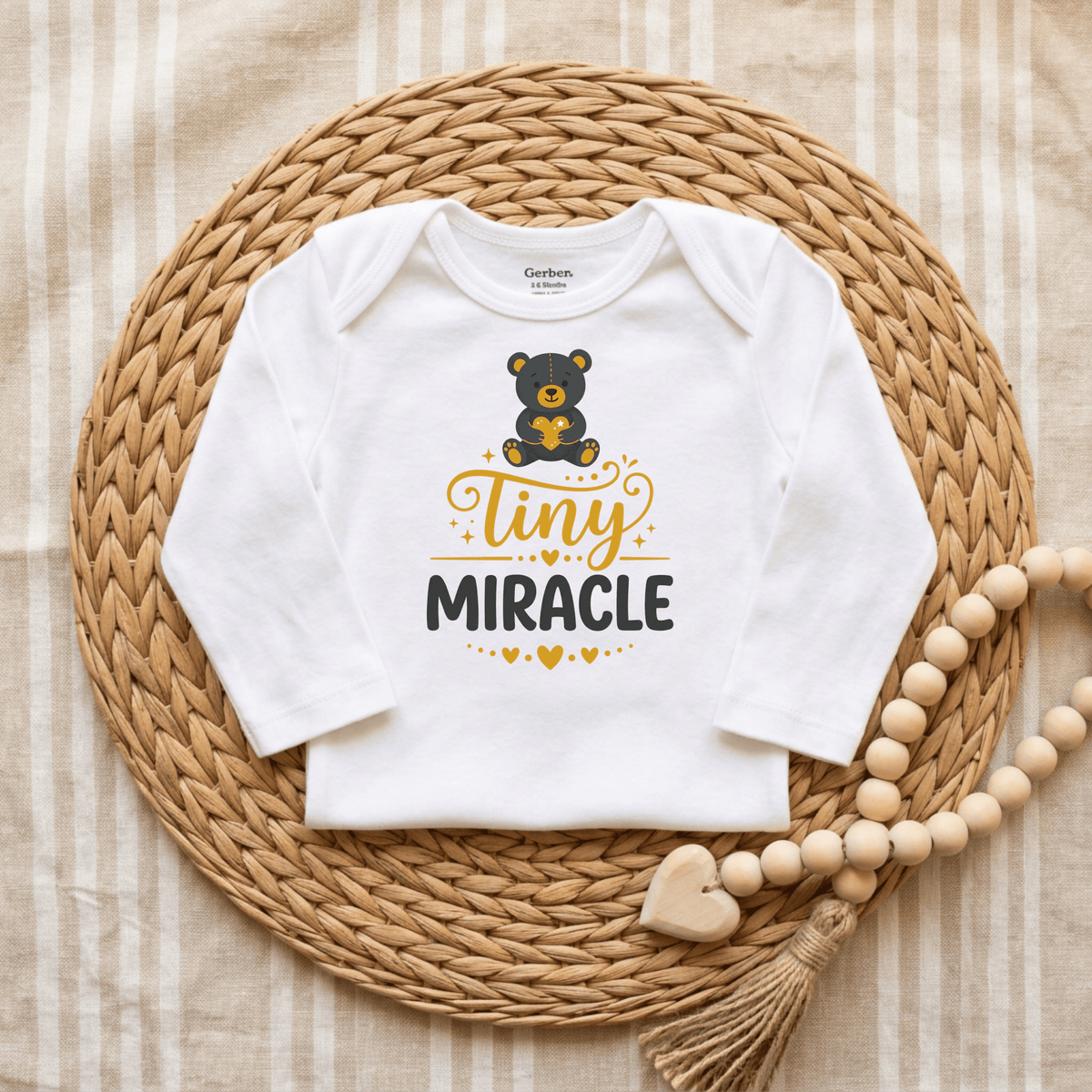The Toodleroos Tiny Miracle Onesie – Cute Baby Teddy Bear Bodysuit is shown on a woven placemat next to wooden beads with a heart charm, all set against a striped fabric background. Featuring an adorable bear graphic and the words “Tiny Miracle,” this white baby onesie makes a perfect newborn bodysuit and thoughtful NICU graduate gift to celebrate your little one’s journey.