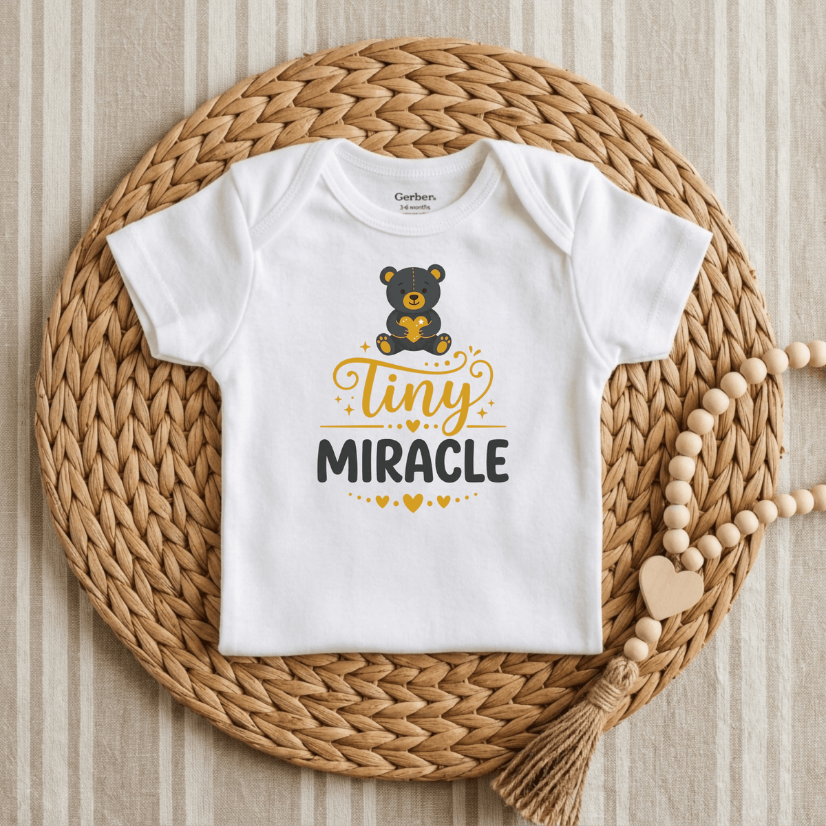 The Toodleroos Tiny Miracle Onesie – Cute Baby Teddy Bear Bodysuit in white, adorned with an adorable bear graphic and the phrase “Tiny Miracle,” is showcased on a woven placemat beside a wooden beaded garland atop striped fabric—making it the perfect gift for a NICU graduate.