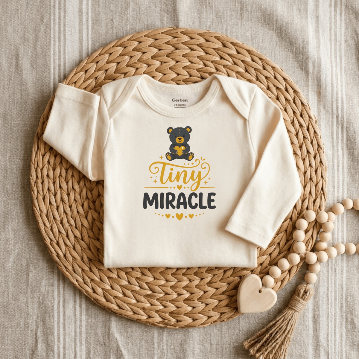 The Toodleroos Tiny Miracle Onesie – Cute Baby Teddy Bear Bodysuit is a cream-colored newborn outfit featuring an adorable bear graphic and the words “tiny MIRACLE” in cheerful yellow and black, shown on a woven placemat beside a wooden beaded toy and heart atop neutral striped fabric—an ideal gift for NICU graduates or anyone looking for cute baby onesies.