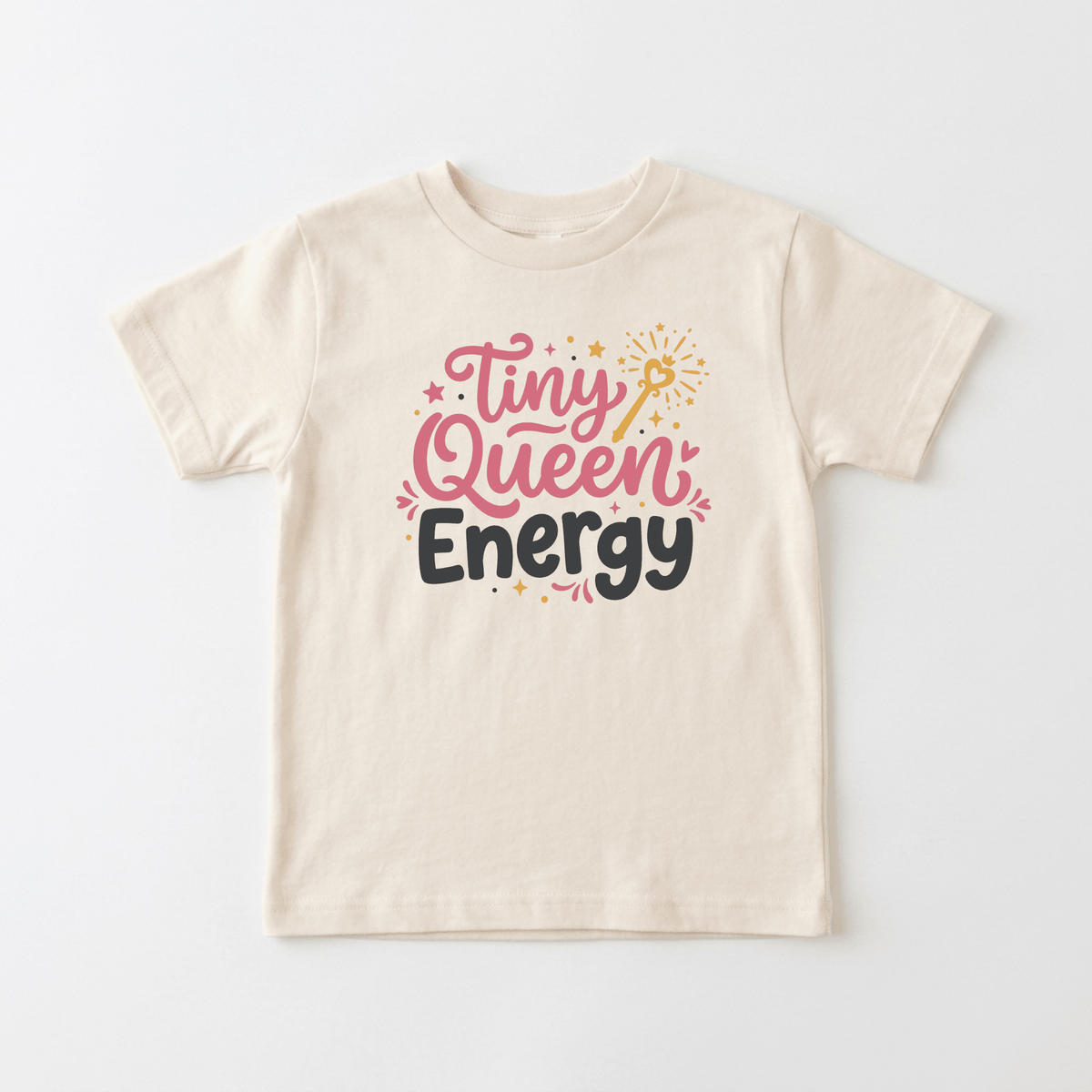 The Toodleroos Tiny Queen Energy Toddler Tee is a cute girls shirt in beige, featuring “Tiny Queen Energy” in pink and black text with star and wand accents—a charming addition to any little one’s wardrobe.