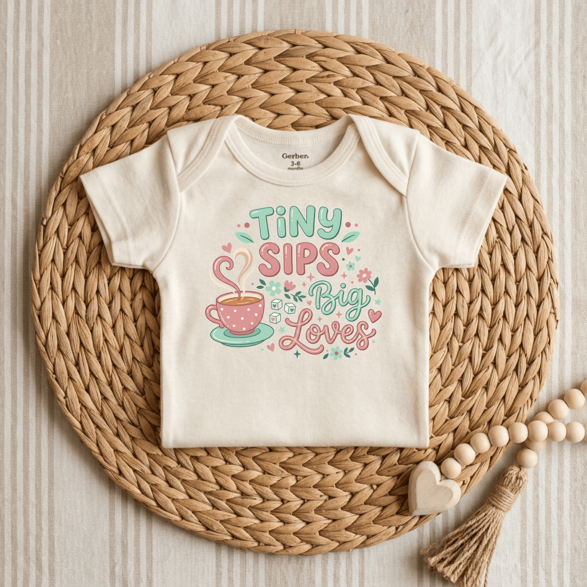 A Toodleroos Tiny Sips Big Loves Onesie, in a soft cream hue, is displayed on a round woven mat. Ideal as a tea party baby onesie, this adorable bodysuit showcases a pink polka-dot teacup graphic with heart-shaped steam and floral accents, alongside the cheerful text “TINY SIPS Big Loves.” Wooden beads placed nearby add to the sweet and whimsical appeal of this cute baby girl teacup bodysuit.