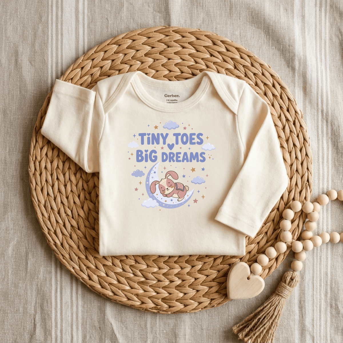 A cream gender-neutral baby bodysuit from Toodleroos, the Tiny Toes Big Dreams Onesie – Cute Sleepy Bunny Bodysuit, is displayed on a woven mat. The outfit features an adorable bunny resting on a moon with the phrase “Tiny Toes Big Dreams,” surrounded by stars and clouds. Ideal for those searching for animal-themed onesies, this charming piece is accented with a wooden bead and heart decoration beside it.