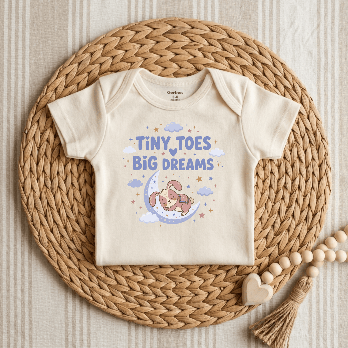 The Toodleroos Tiny Toes Big Dreams Onesie – Cute Sleepy Bunny Bodysuit features a cream color and a charming design of a sleeping bunny resting on a crescent moon with clouds, stars, and the phrase “Tiny Toes Big Dreams.” Displayed on a woven placemat beside a beaded garland, this sweet gender-neutral onesie brings whimsical charm to any nursery collection.