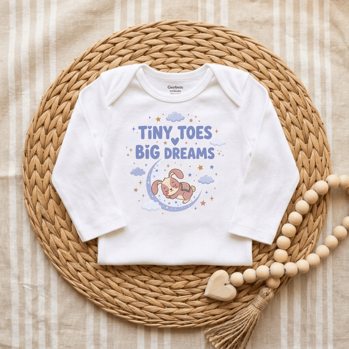 A white baby onesie by Toodleroos is displayed on a woven mat with wooden beads. The gender-neutral “Tiny Toes Big Dreams Onesie – Cute Sleepy Bunny Bodysuit” features a sleeping bunny on a crescent moon surrounded by clouds and stars in soft pastel colors—perfect for those who love sweet animal-themed bodysuits.