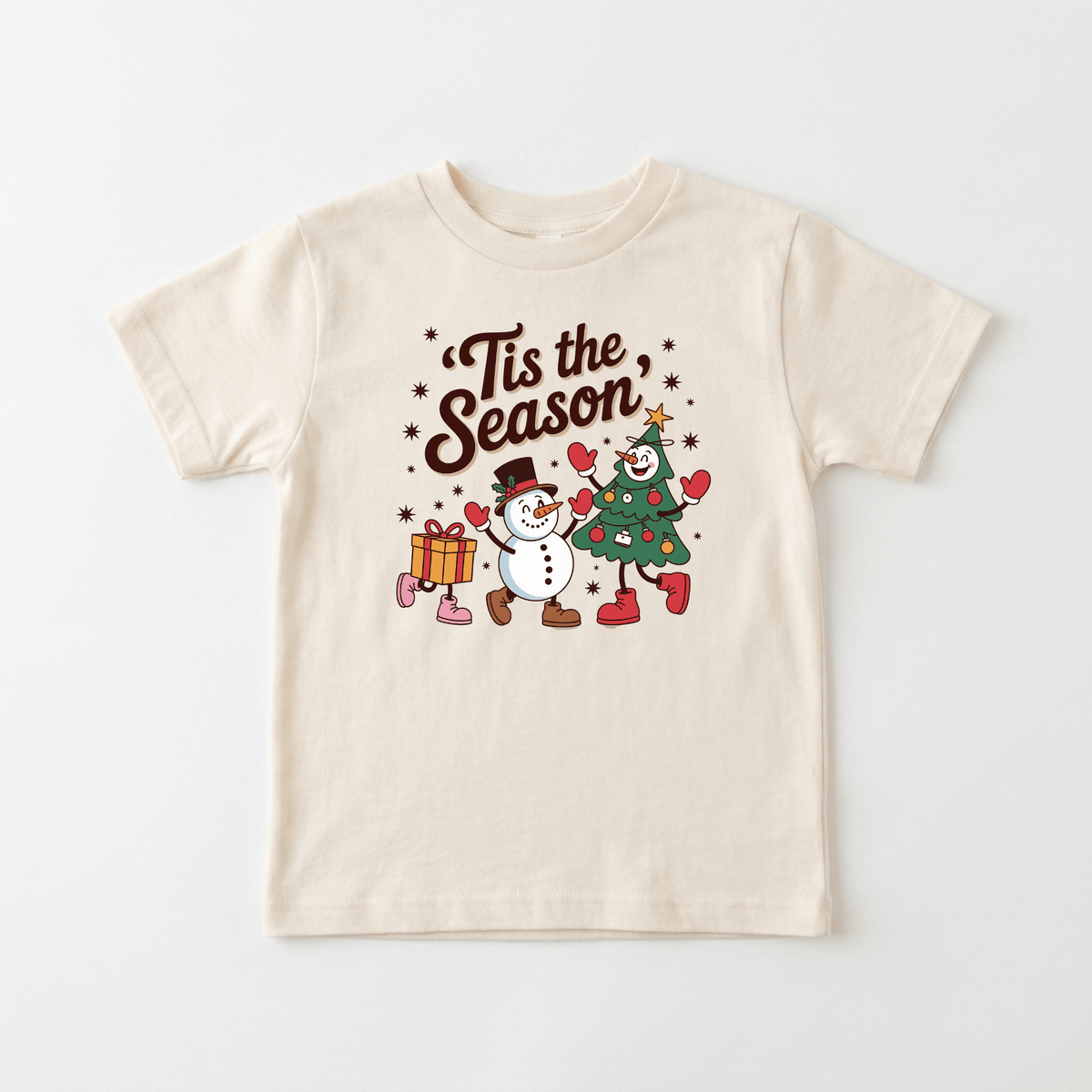 The Toodleroos ’Tis the Season Toddler Tee is a cream-colored, retro Christmas kids shirt featuring a festive design with a snowman, Christmas tree, and gift box holding hands beneath the phrase “Tis the Season,” surrounded by stars and hearts—perfect for adding vintage holiday cheer to your toddler’s wardrobe.