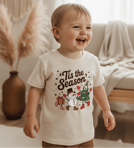 A smiling toddler wears the Toodleroos ’Tis the Season Toddler Tee – a retro Christmas kids shirt with festive graphics – while standing indoors surrounded by neutral decor and soft natural light.