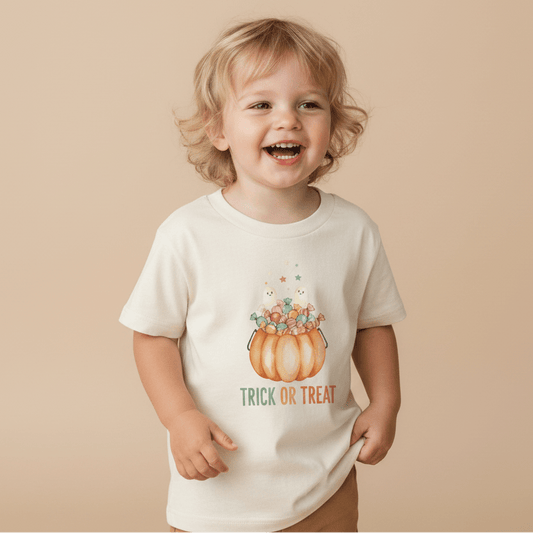 A happy child with wavy blonde hair wears the Toodleroos Toddler Halloween Shirt – Trick or Treat Pumpkin & Ghost Tee, showcasing a festive pumpkin overflowing with candy and a playful “Trick or Treat” design, set against a beige background.