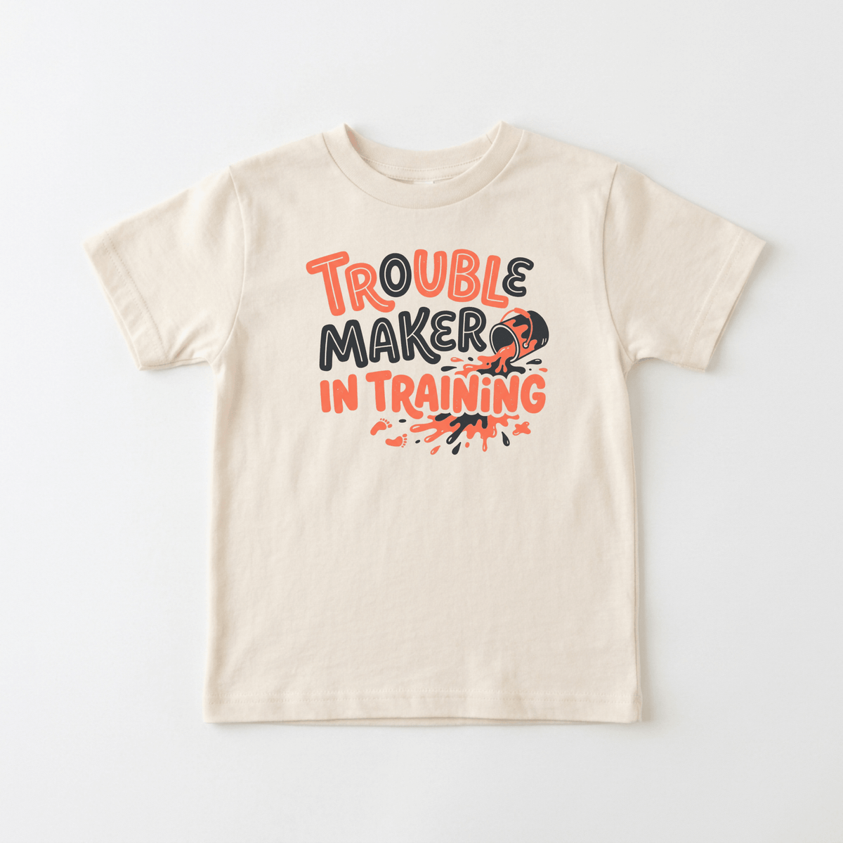 The Toodleroos Trouble Maker in Training Toddler Shirt is a funny toddler tee in beige, showcasing bold black and orange text that reads “Trouble Maker in Training,” along with a small child silhouette and lively splatter graphics. This playful kids shirt is displayed flat against a white background.