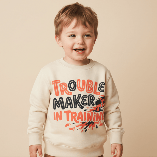 A smiling young child wears the Toodleroos Trouble Maker in Training Toddler Sweatshirt – a cozy cream fleece pullover featuring playful red, black, and orange lettering and designs—perfect as an adorable toddler gift idea.