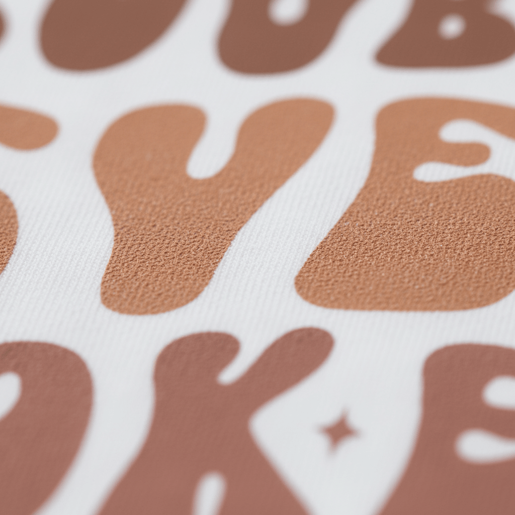 A close-up of the Toodleroos “Trouble Never Looked So Sweet Kids Shirt – Funny Retro Tee” shows large, textured brown and tan bubble letters spelling BYE in a playful retro style on soft white fabric—an adorable and humorous kids’ shirt that truly captures the brand’s sweet approach to mischief.