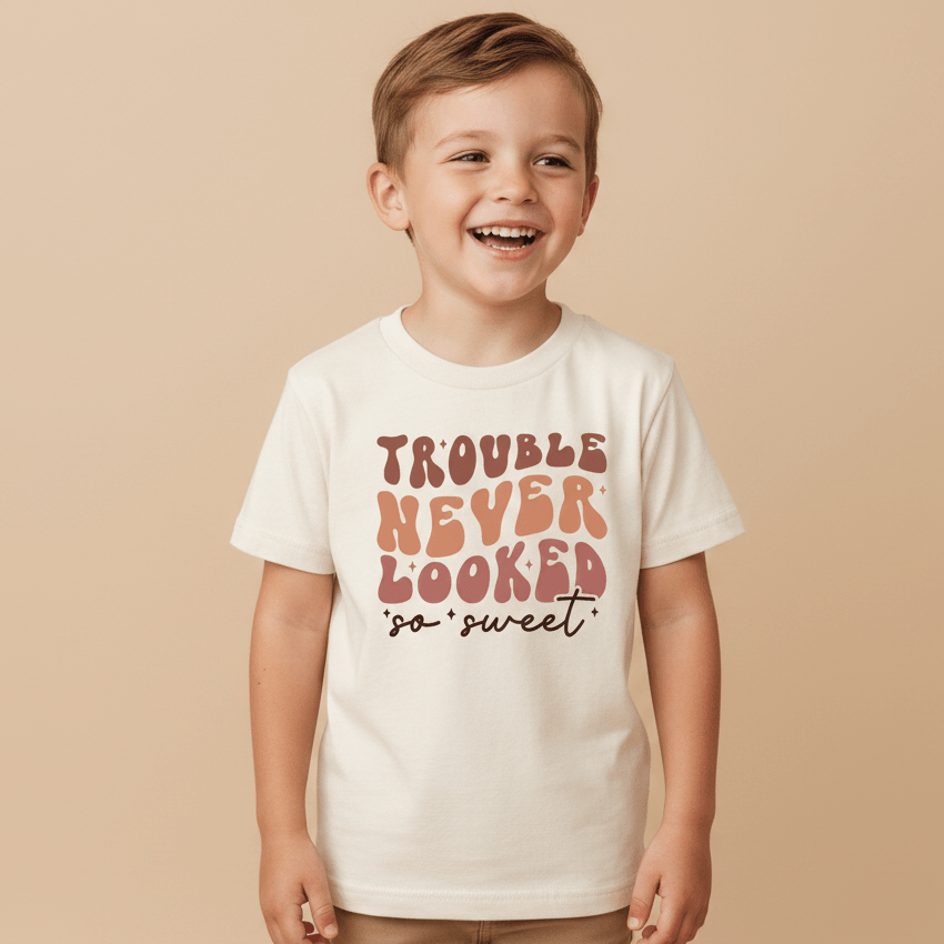 A cheerful young boy wearing the Toodleroos “Trouble Never Looked So Sweet” Kids Shirt—a funny retro white tee with colorful text—stands against a beige background.