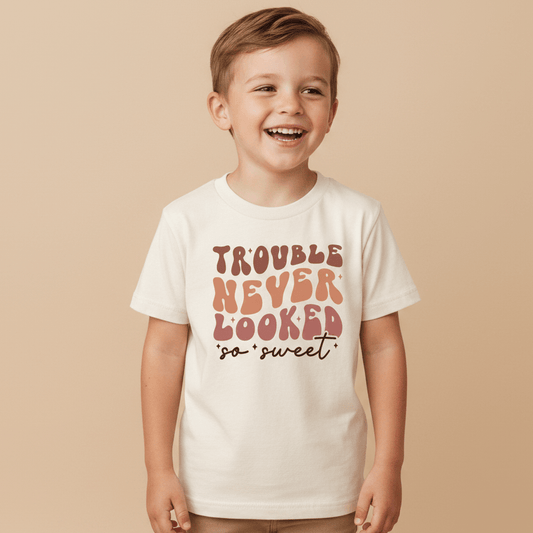 A cheerful young boy wearing the Toodleroos “Trouble Never Looked So Sweet” Kids Shirt—a funny retro white tee with colorful text—stands against a beige background.