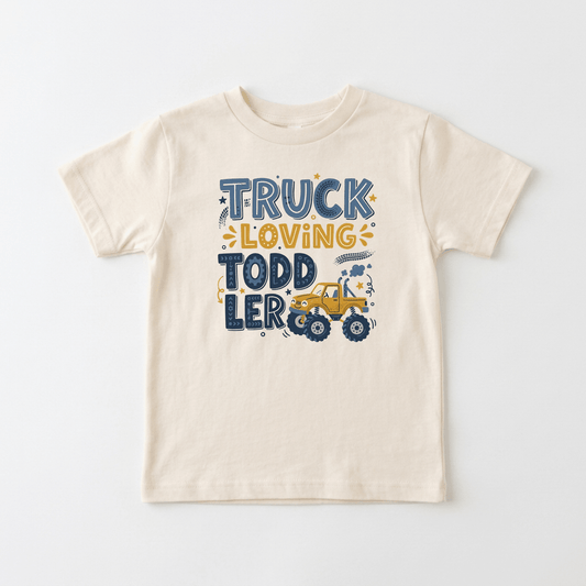 The Toodleroos Truck Loving Toddler Boys Shirt – Monster Truck Tee is a cream-colored t-shirt featuring the phrase “Truck Loving Toddler” in blue and yellow, a playful cartoon yellow truck, and decorative stars and dots—making it an ideal addition to boys shirt collections or as a fun monster truck shirt for your little one.