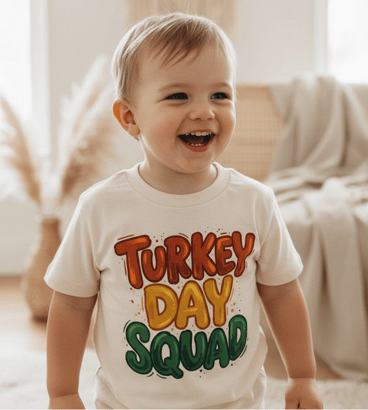 A happy toddler wears the Toodleroos Turkey Day Squad Toddler Tee – Thanksgiving Kids Shirt, standing indoors with neutral decor in the background—an adorable choice for anyone seeking a cute Thanksgiving toddler shirt or festive fall kids shirts.