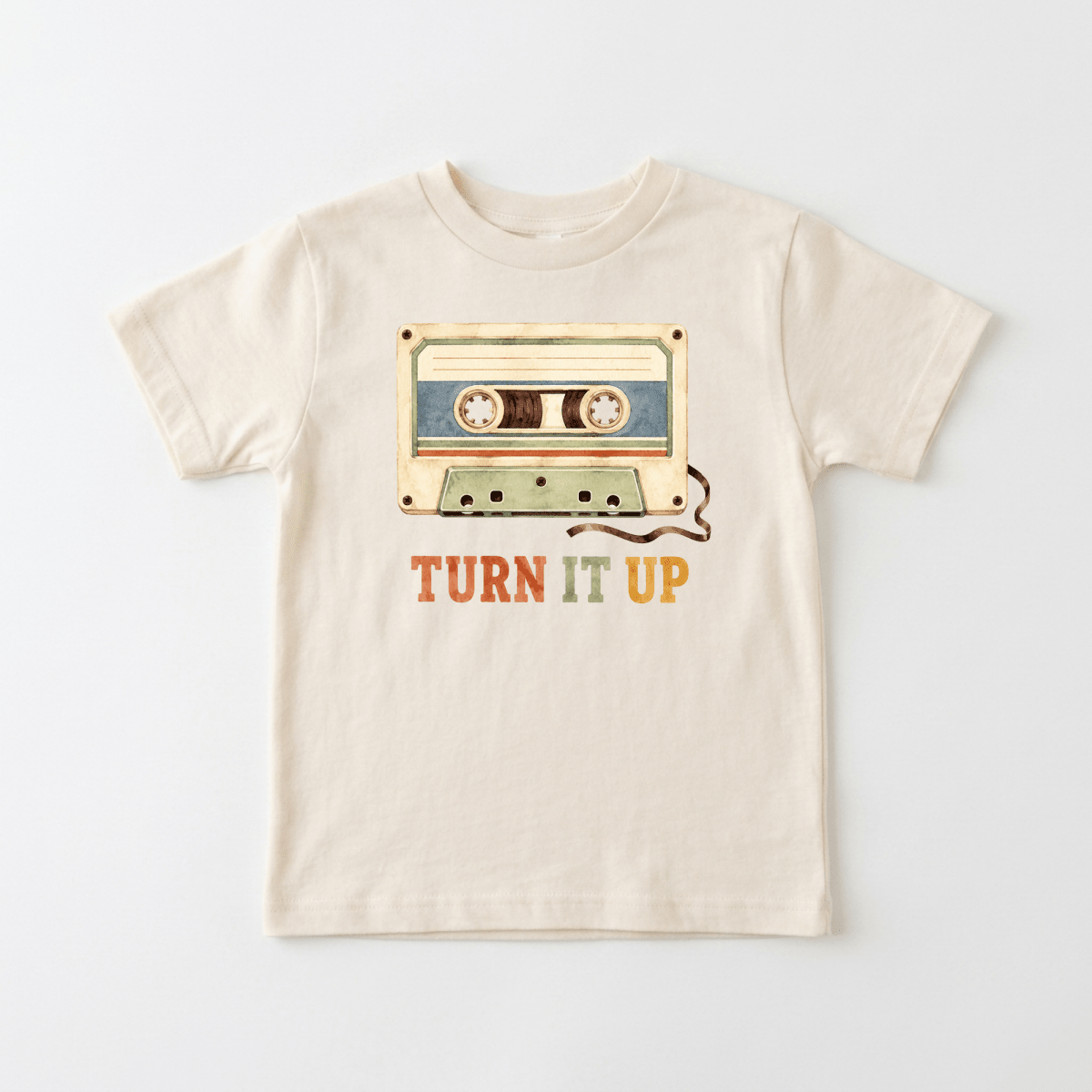The Toodleroos Toddler Music Shirt – Turn It Up Retro Cassette Graphic Tee is a beige t-shirt showcasing a vintage cassette tape image above the phrase “TURN IT UP” in red, orange, and green lettering—a perfect retro cassette shirt and graphic tee for music-loving kids.