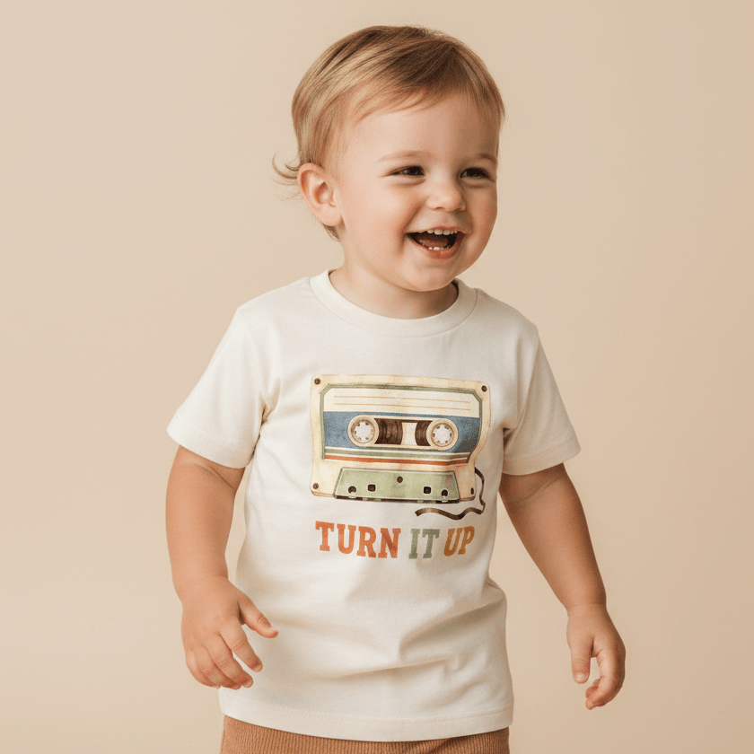 A cheerful toddler stands against a plain beige background wearing the Toodleroos Toddler Music Shirt – Turn It Up Retro Cassette Graphic Tee, a white t-shirt showcasing a vibrant cassette tape graphic and the phrase “TURN IT UP” on the front.