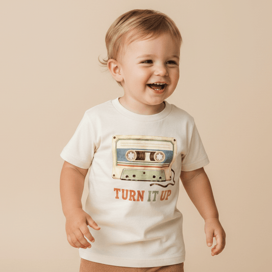 A cheerful toddler stands against a plain beige background wearing the Toodleroos Toddler Music Shirt – Turn It Up Retro Cassette Graphic Tee, a white t-shirt showcasing a vibrant cassette tape graphic and the phrase “TURN IT UP” on the front.