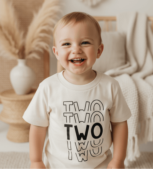 A happy toddler with short hair is wearing the Toodleroos Two Birthday Toddler Shirt – a gender-neutral 2nd Birthday Tee featuring the word “TWO” repeated three times – while standing in a cozy, neutral-toned room adorned with textured blankets and pampas grass.