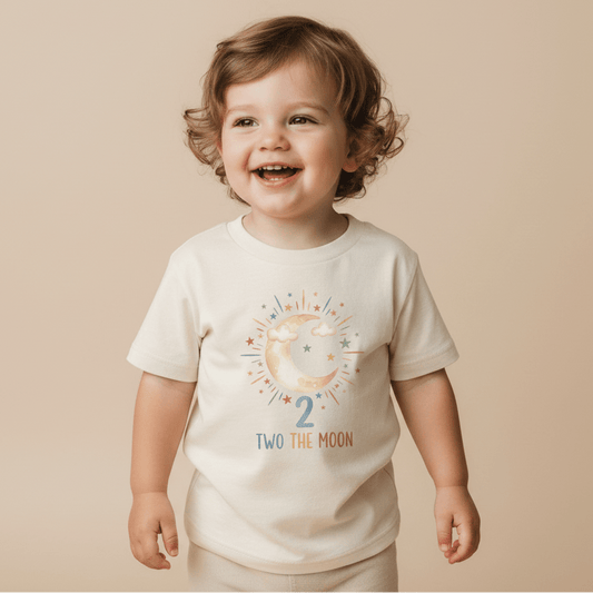 A smiling young child with curly brown hair wears the Toodleroos 2nd Birthday Toddler Shirt – Two the Moon Celestial Short-Sleeve Tee, featuring a moon, stars, the number 2, and “Two the Moon,” while standing against a neutral background.