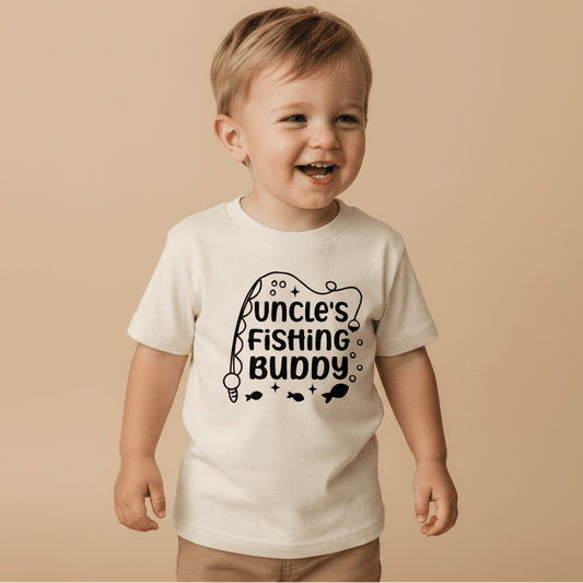 A cheerful toddler stands against a beige background, wearing the Toodleroos Uncle’s Fishing Buddy Toddler Boys Shirt—a cream tee featuring playful fishing-themed graphics and “Uncle’s Fishing Buddy” text. This adorable fishing rod tee is a perfect Father’s Day gift or a fun surprise for your favorite uncle.