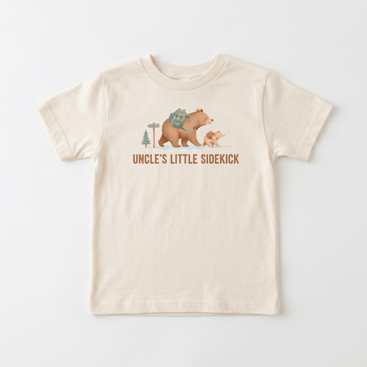 The Toodleroos Uncle’s Little Sidekick Toddler Shirt – Hiking Bear Tee is a beige t-shirt displaying an illustration of a large bear with a backpack and a small bear cub by a pine tree and sign, with “UNCLE’S LITTLE SIDEKICK” printed below—making it the perfect uncle toddler shirt or hiking bear toddler tee for outdoor adventures together.
