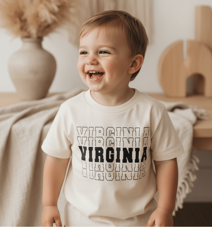 A smiling toddler stands indoors by a table with neutral decor, including a beige vase filled with pampas grass and a draped cloth, proudly wearing the Toodleroos Virginia Varsity Toddler Shirt – Kids State Pride Tee featuring a varsity-inspired graphic.