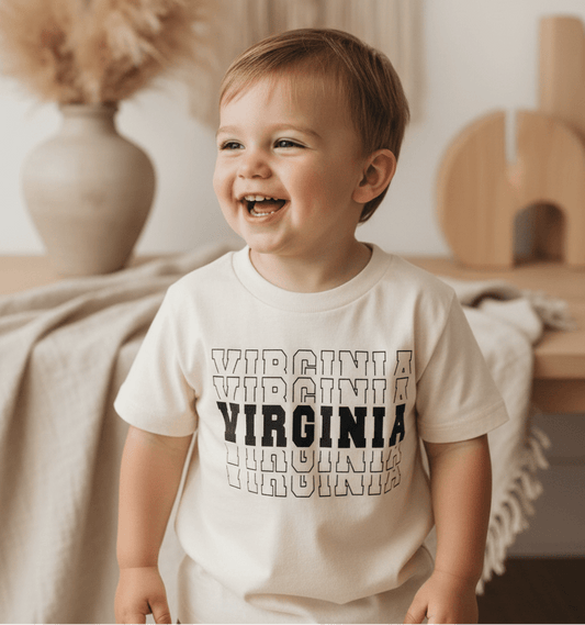A smiling toddler stands indoors by a table with neutral decor, including a beige vase filled with pampas grass and a draped cloth, proudly wearing the Toodleroos Virginia Varsity Toddler Shirt – Kids State Pride Tee featuring a varsity-inspired graphic.