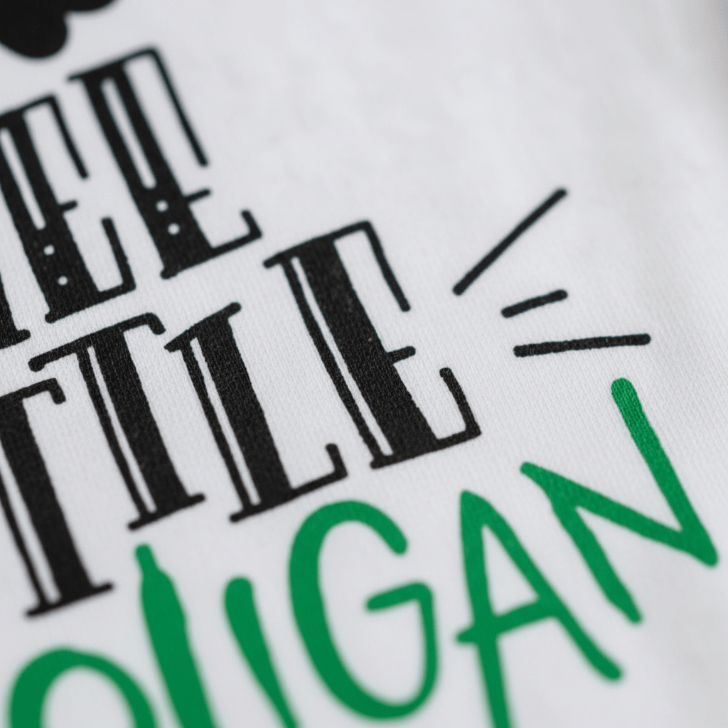 Close-up of the Wee Little Hooligan Baby Onesie by Toodleroos, featuring bold black and green text in various fonts. The words “LITTLE” and part of “HOOLIGAN” stand out with decorative lines accenting the playful design—making this St. Patrick’s Day bodysuit an adorable choice for your little one’s first celebration. A truly funny baby shower gift, this Toodleroos St. Patrick’s Day onesie brings a joyful spirit to any festivity.