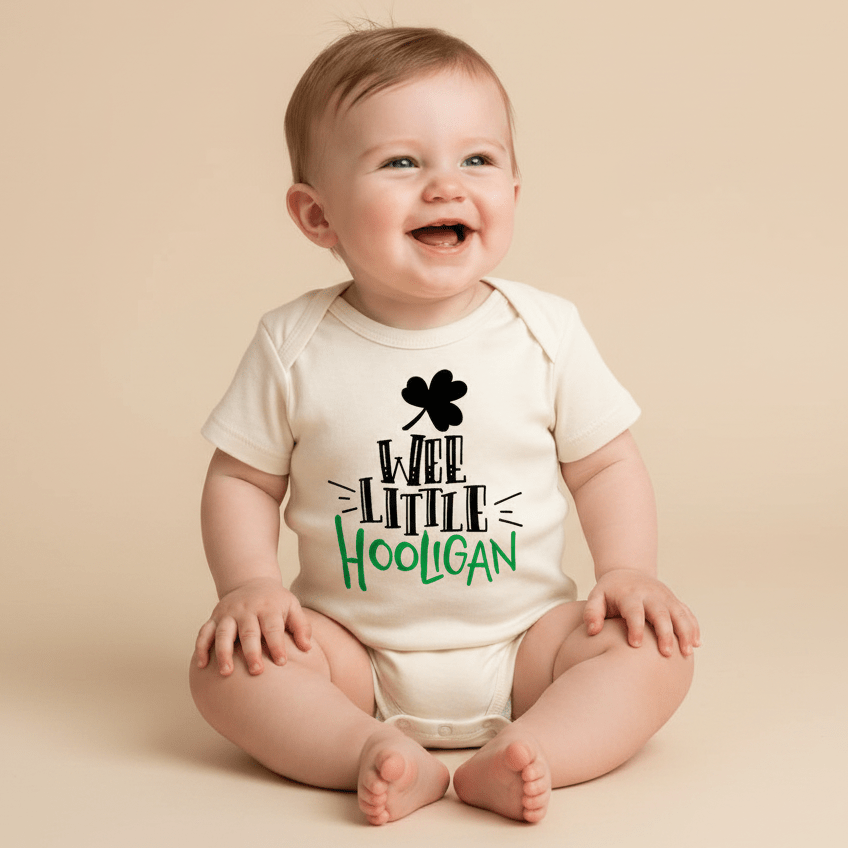 A happy baby sits on a beige surface, dressed in the Toodleroos “Wee Little Hooligan Baby Onesie – St. Patrick’s Day Bodysuit,” featuring cream fabric with “Wee Little Hooligan” printed in playful black and green letters—an amusing baby shower gift ideal for celebrating St. Patrick’s Day.