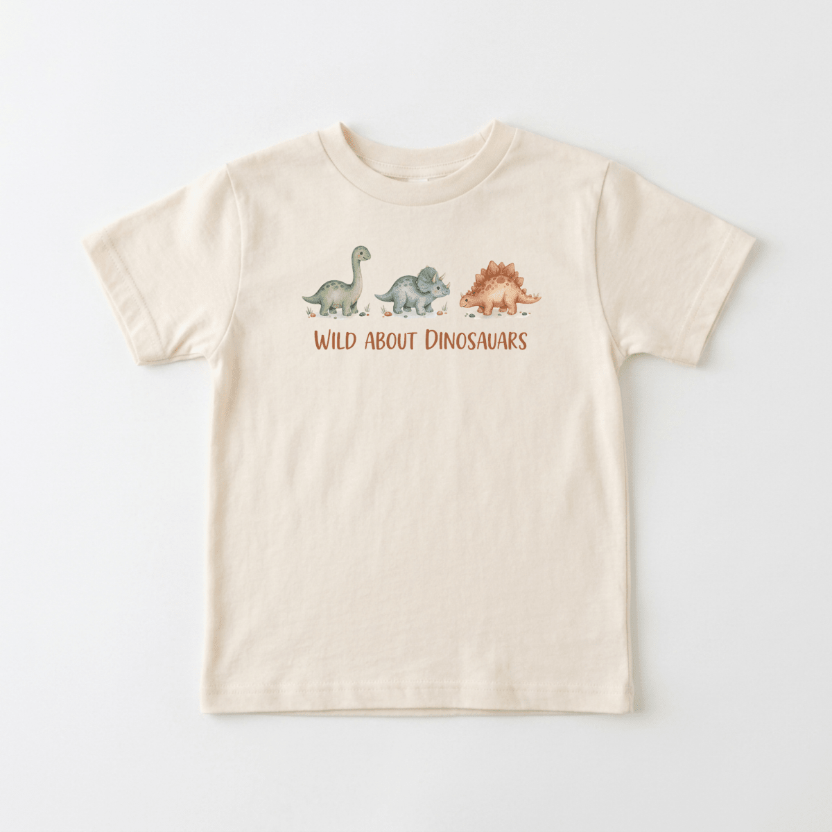 Dress your little one in the Toodleroos Wild About Dinosaurs Toddler Shirt – Watercolor Dino Tee, a beige graphic tee featuring watercolor illustrations of three dinosaurs and bold orange “WILD ABOUT DINOSAURS” lettering above them. This playful toddler shirt is perfect for kids who love prehistoric adventures.
