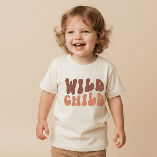 A smiling toddler with curly hair wears the Toodleroos Wild Child Kids Shirt – Boho Retro Tee, featuring bold, retro-style lettering in cream against a plain beige background—an adorable tribute to vintage-inspired kids’ fashion.