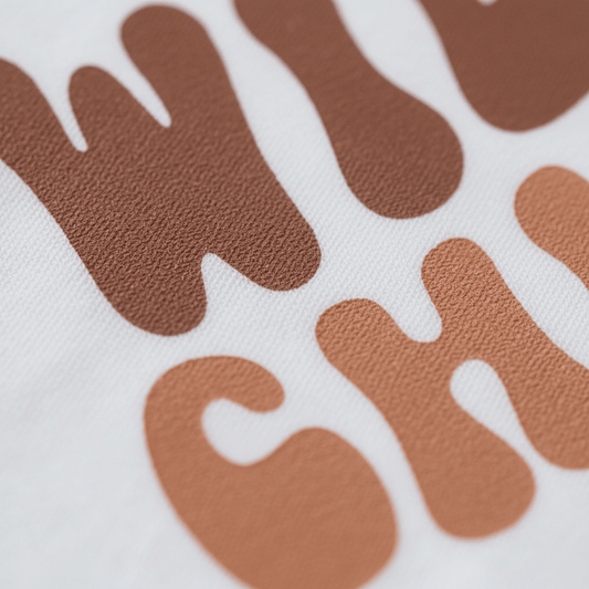 Close-up of brown, wavy, textured letters on a white fabric background, partially displaying “WI” and “CH.” The bold screen-printed lettering channels the playful spirit of the Toodleroos Wild Child Kids Shirt – Boho Retro Tee, adding a touch of vintage flair inspired by retro children’s styles.