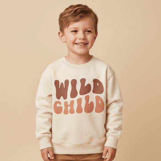 A smiling young boy wearing light brown pants and the Toodleroos Wild Child Kids Sweatshirt – a cozy, boho retro pullover featuring bold, retro-style “WILD CHILD” lettering – stands against a beige background.