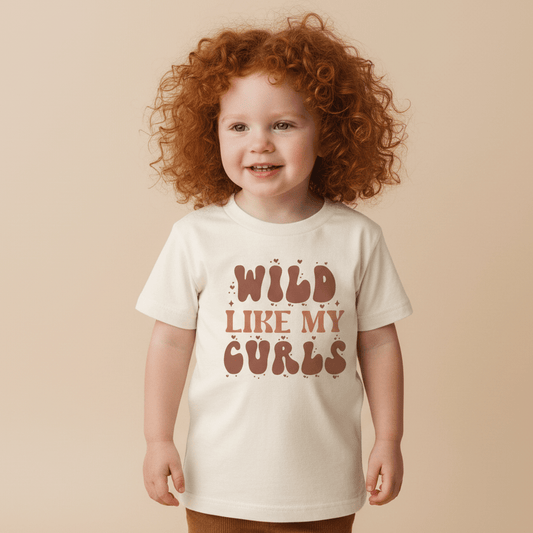 A smiling young child with curly red hair wears the Toodleroos “Wild Like My Curls” Kids Shirt – a boho retro tee featuring bold, playful brown lettering—standing against a plain beige background.