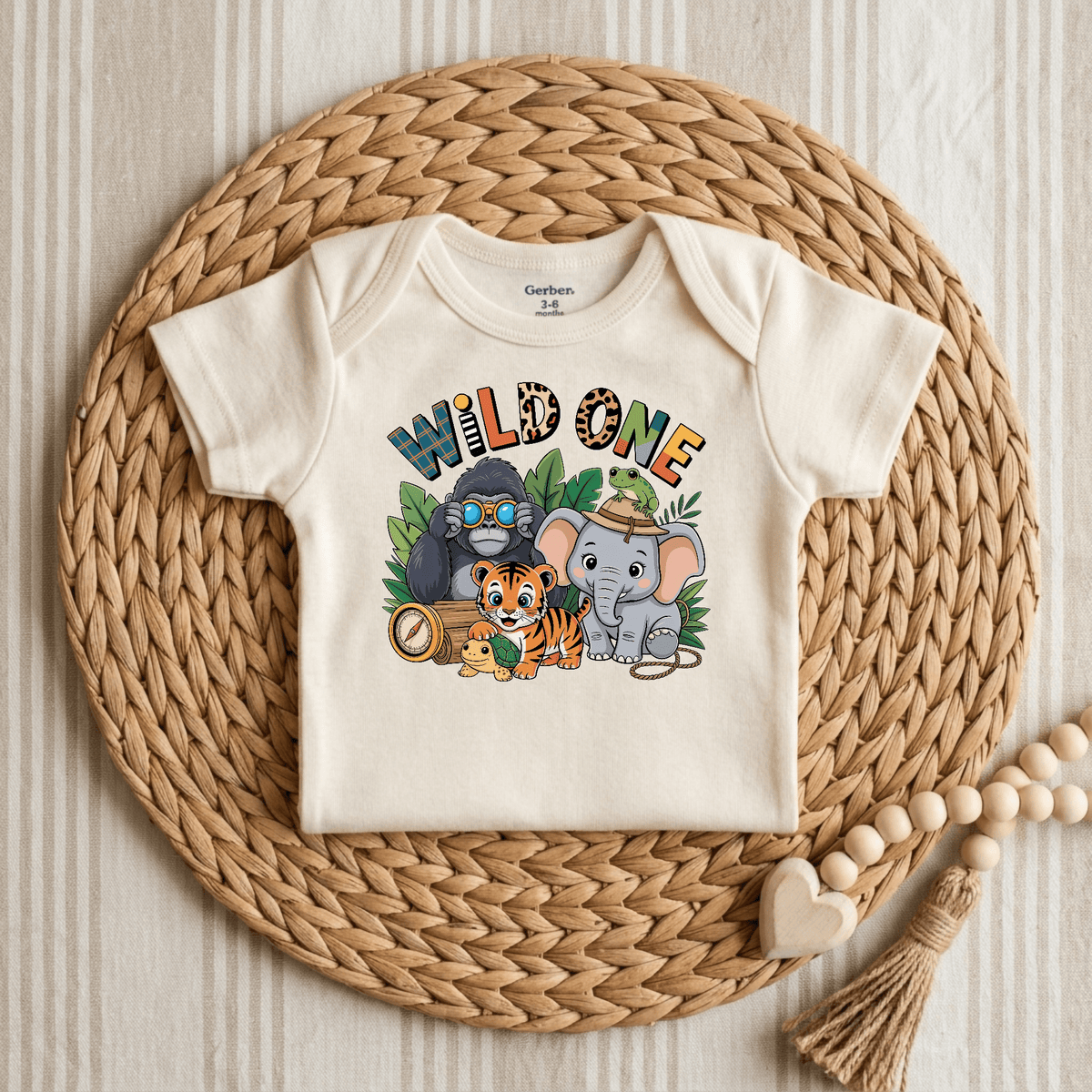 A cream-colored Toodleroos Wild One 1st Birthday Baby Onesie – Jungle Safari Bodysuit, labeled 3-6 months, is displayed on a round woven placemat. The design features the words “WILD ONE” in safari-themed fonts above playful illustrations of a gorilla with binoculars, a tiger, an elephant wearing a hat with a frog, and a small turtle holding a compass—making it the perfect jungle animal bodysuit for your little explorer’s first birthday safari adventure.