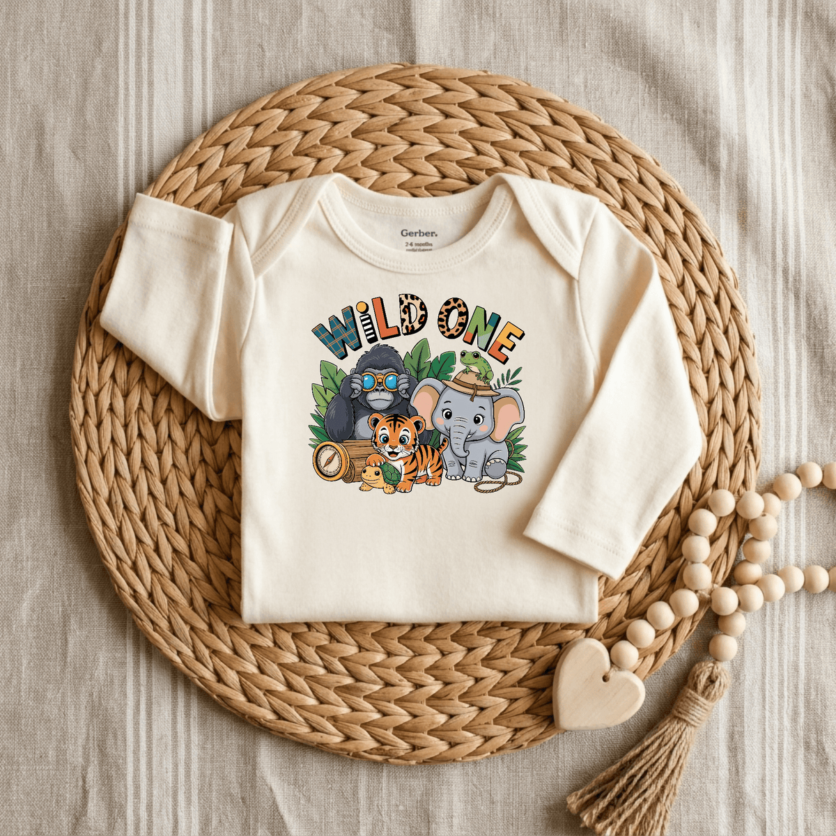 A cream-colored, long-sleeve Toodleroos Wild One 1st Birthday Baby Onesie – Jungle Safari Bodysuit is centered on a woven placemat. This adorable onesie features a playful graphic of a gorilla, tiger, elephant, and turtle with the text “WILD ONE” above them—making it perfect for celebrating a safari-themed first birthday. Wood beads rest to the side on a striped fabric background.