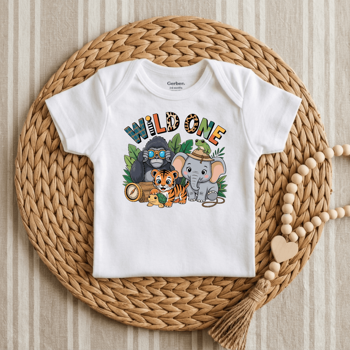 The Toodleroos Wild One 1st Birthday Baby Onesie – Jungle Safari Bodysuit, featuring playful “Wild One” text and charming cartoon animals (gorilla, tiger, elephant, zebra) on white fabric, is displayed on a woven placemat next to a heart-accented wooden bead garland. Set against a beige striped background, this adorable onesie is perfect for your little one’s safari-themed first birthday celebration.